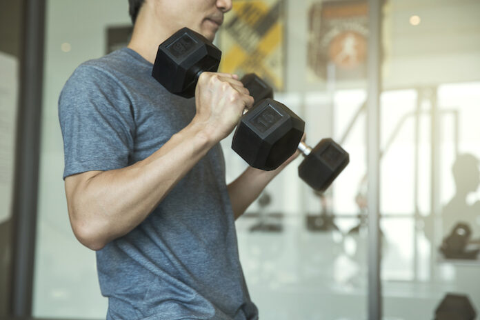 Young man beginner exercising with dumbbell flexing muscles at gym, sport training concept